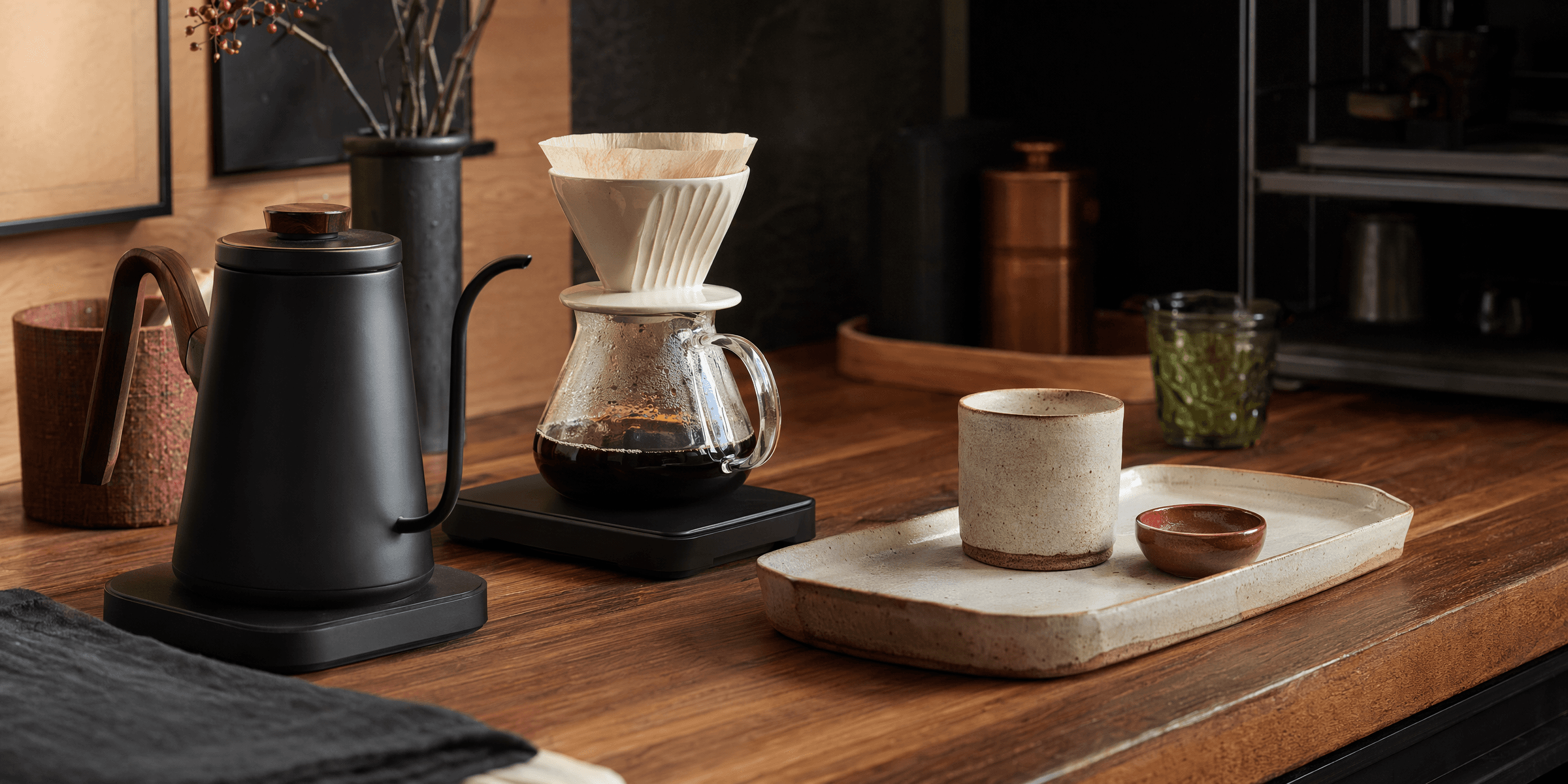 A warm coffee and tea counter with a pour-over kettle, ceramic dripper, glass server and tray on a wooden worktop